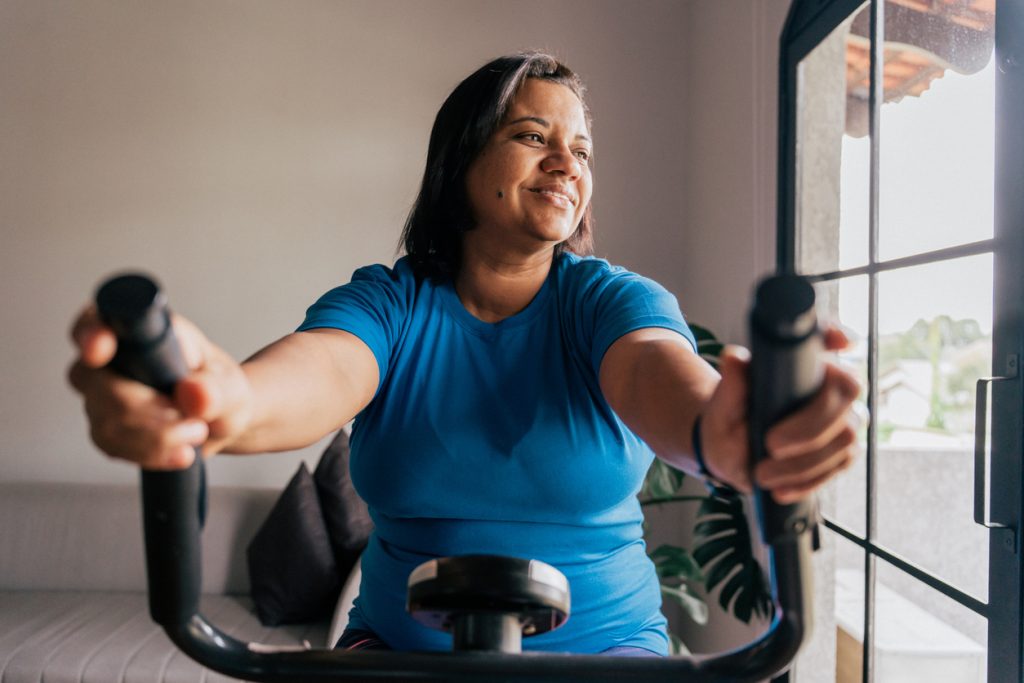 Mulher sorridente e motivada praticando exercícios físicos em uma bicicleta ergométrica dentro de casa, representando a importância do movimento e do estilo de vida saudável como um dos principais tratamentos para resistência à insulina.