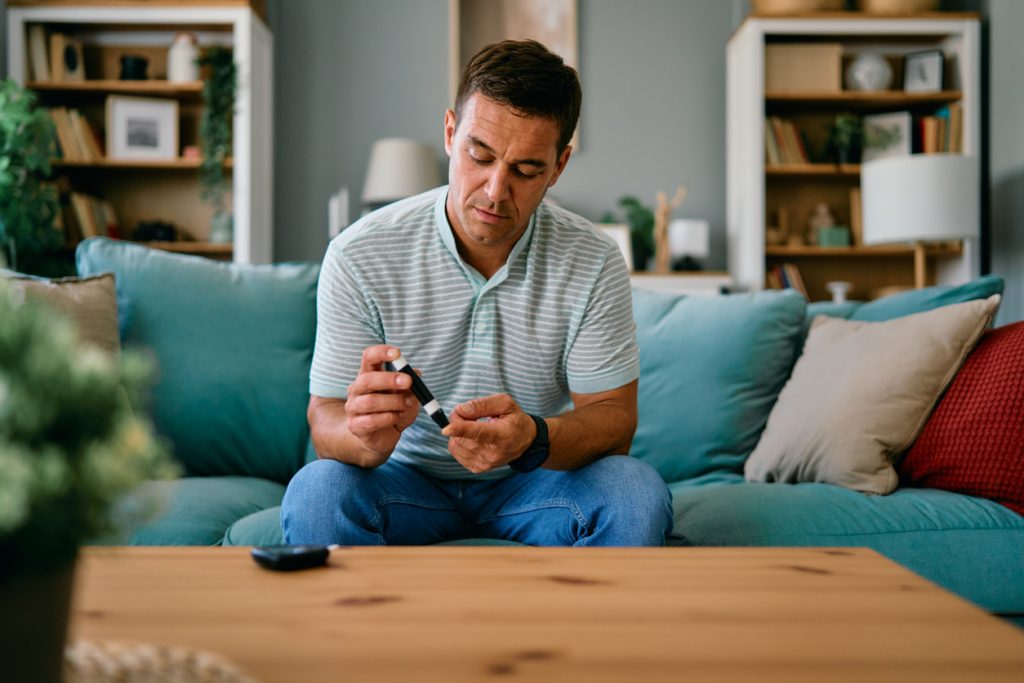 Homem adulto sentado no sofá de sua sala usando um aparelho glicosímetro na ponta do dedo para medir os níveis de açúcar no sangue, ilustrando o monitoramento necessário para entender as diferenças entre diabetes e resistência à insulina.