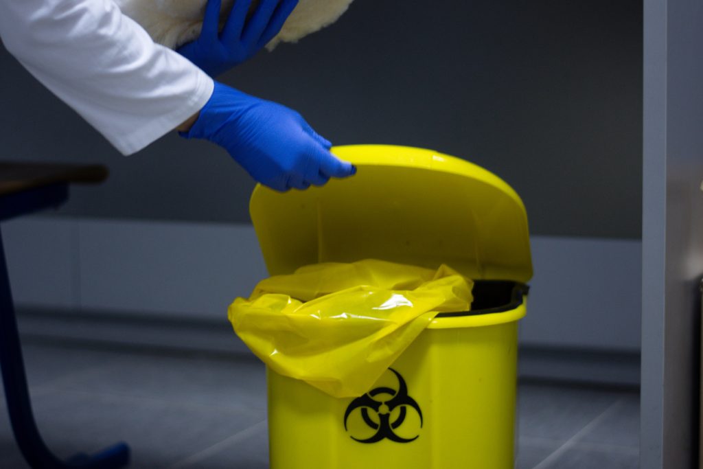 Mãos com luvas azuis e braços com mangas de jaleco abrindo lixo hospitalar.