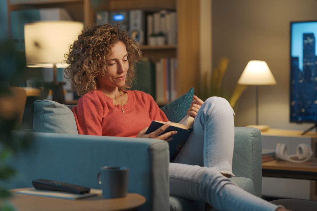 Mulher sentada confortavelmente em uma poltrona à noite, iluminada pela luz suave e amarelada de um abajur, lendo um livro, exemplificando uma excelente prática de higiene do sono para desacelerar o corpo e a mente.