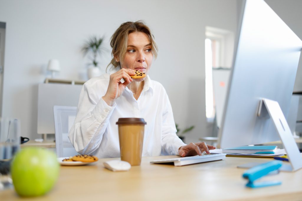 Mulher trabalhando em frente a um monitor no escritório, segurando um copo de café e comendo um biscoito com gotas de chocolate, ilustrando como o excesso de cafeína e açúcar pode impactar negativamente a higiene do sono.