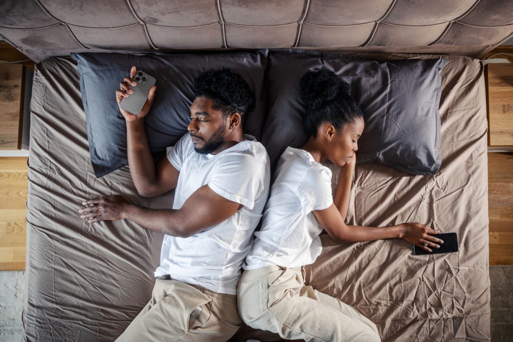 Vista superior de um casal deitado na cama, dormindo de costas um para o outro, com ambos ainda segurando seus smartphones nas mãos, destacando um dos erros mais comuns que prejudicam a higiene do sono.