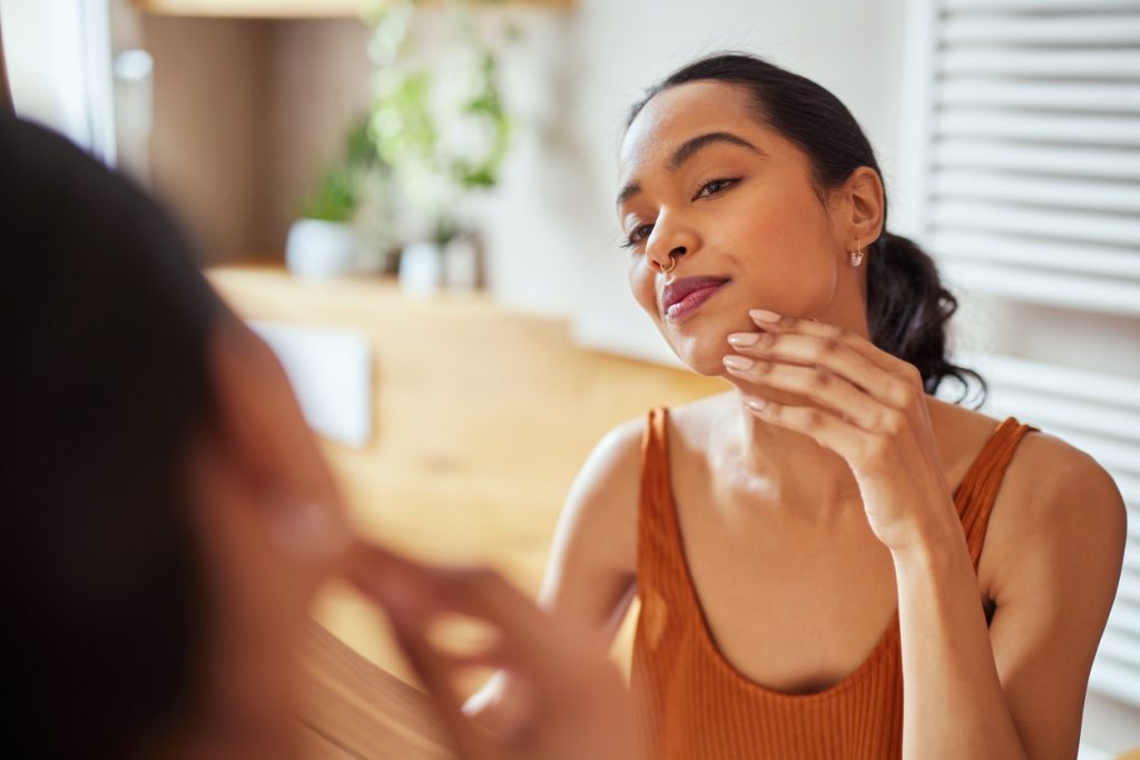 Mulher jovem com piercing no nariz e unhas claras se observa no espelho, passando a mão na bochecha, demonstrando preocupação com a firmeza da pele e a flacidez facial.