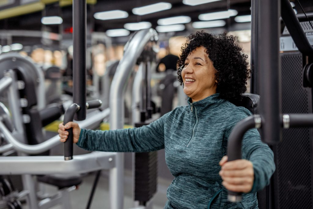 Mulher de meia-idade sorrindo enquanto realiza exercício de força em uma máquina de academia para preservar a massa muscular e a saúde da mulher na menopausa.