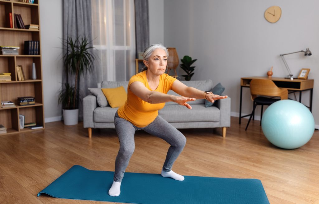 Mulher idosa com roupas de ginástica e cabelo preso fazendo agachamento (peso corporal) no meio de uma sala residencial em cima de um tapete de yoga. Uma ótima opção para incluir na sua rotina de exercícios para fazer em casa.