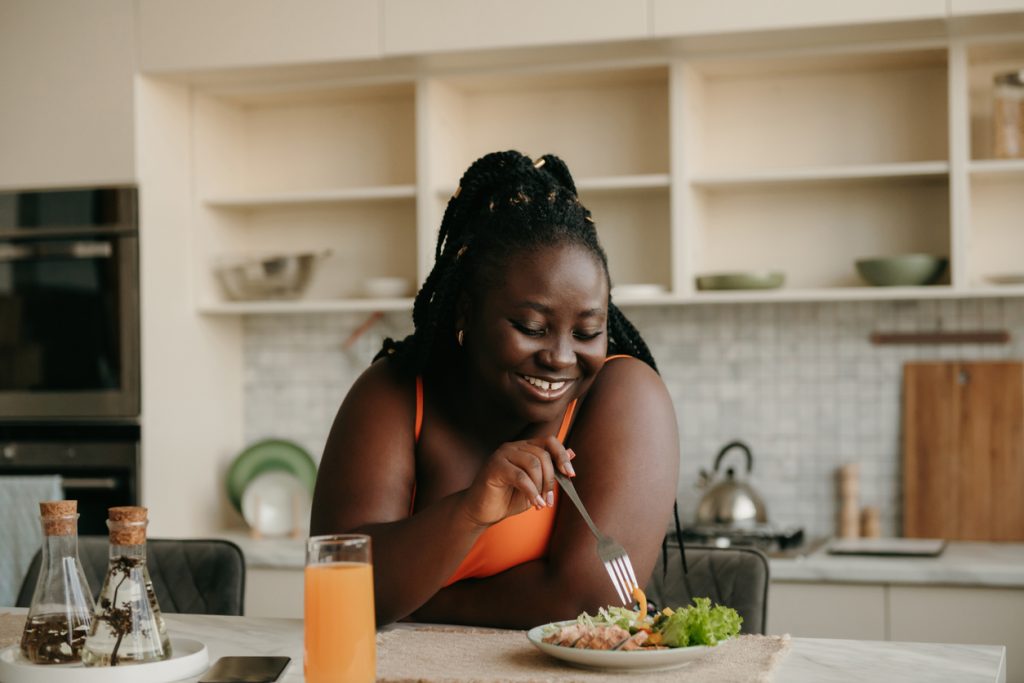 Mulher negra sorrindo enquanto come uma salada nutritiva na cozinha de casa, demonstrando que uma dieta balanceada e saudável é fundamental para quem busca como perder gordura abdominal.