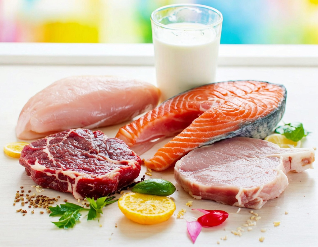 Bancada branca com alguns alimentos ricos em creatina dispostos em evidência - como carne vermelha, peito de frango, carne suína, um pedaço de salmão e copo de leite.