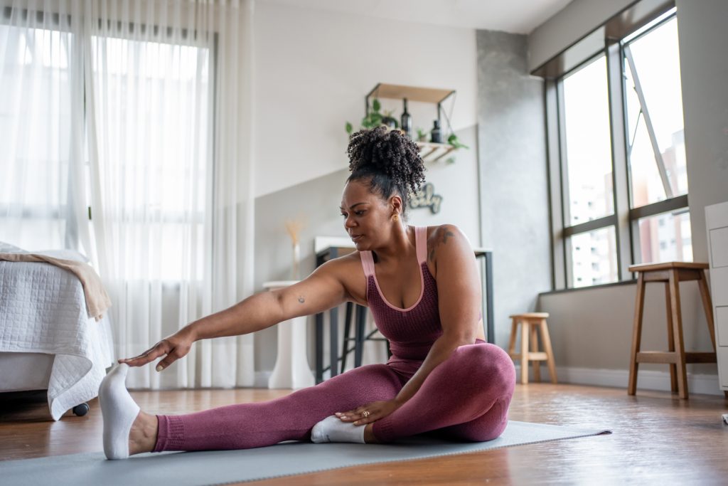 Mulher negra com roupas de ginástica vinho sentada em um tapete na sala, esticando o braço para alcançar a ponta do pé. O alongamento é uma etapa essencial para iniciar ou finalizar sua série de exercícios para fazer em casa com segurança.