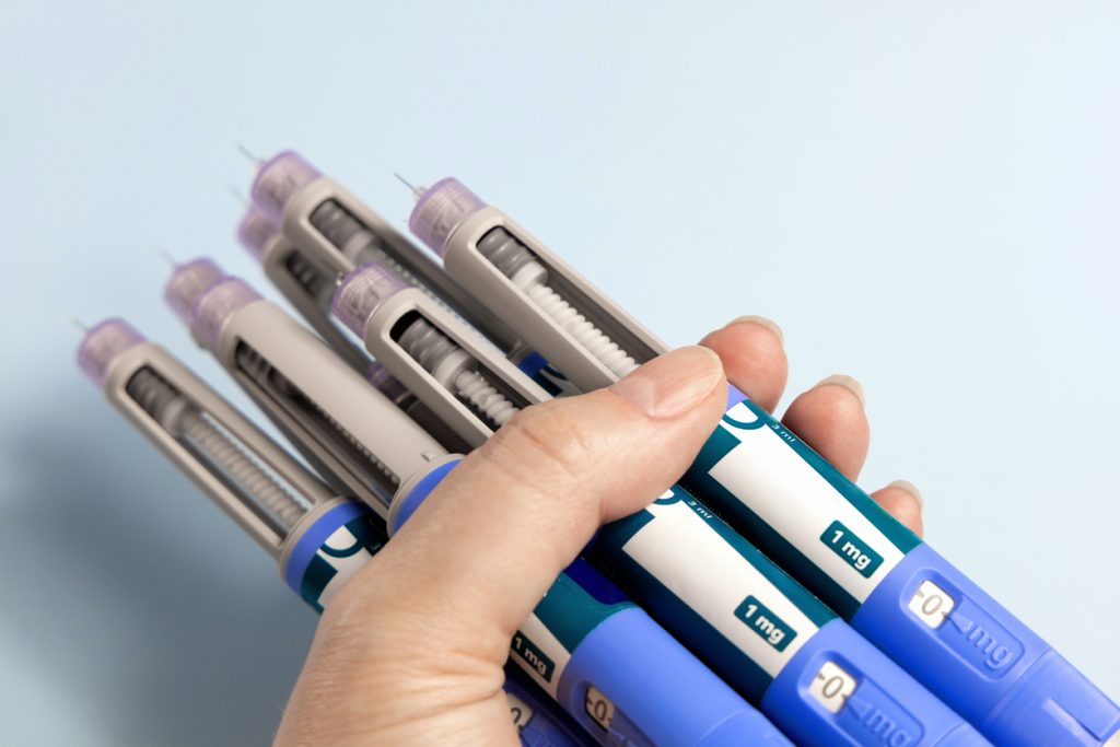 Mão segurando diversas canetas aplicadoras de medicação (análogos de GLP-1) em diferentes dosagens (1 mg), utilizadas no tratamento de diabetes tipo 2 e obesidade.