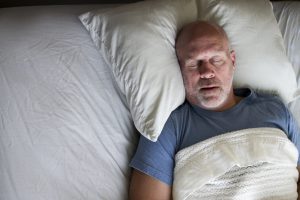 Homem calvo dormindo em uma cama com travesseiros brancos e roupas de cama claras, ilustrando o tema da apneia do sono e a busca pela qualidade do descanso.