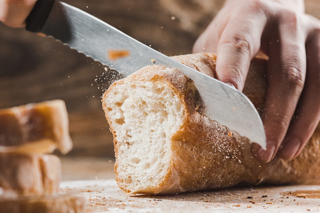 Foco nas mãos de uma pessoa usando uma faca de serra para cortar uma fatia de pão branco de casca crocante sobre uma tábua de madeira, ilustrando as dúvidas comuns sobre o consumo de carboidratos na dieta para diabetes.