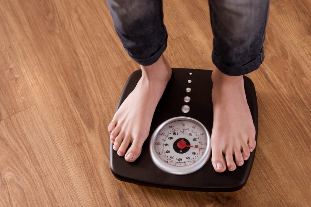 Pés de uma pessoa em pé sobre uma balança analógica de chão, ressaltando a importância do controle do peso corporal para evitar e identificar o pré-diabetes.
