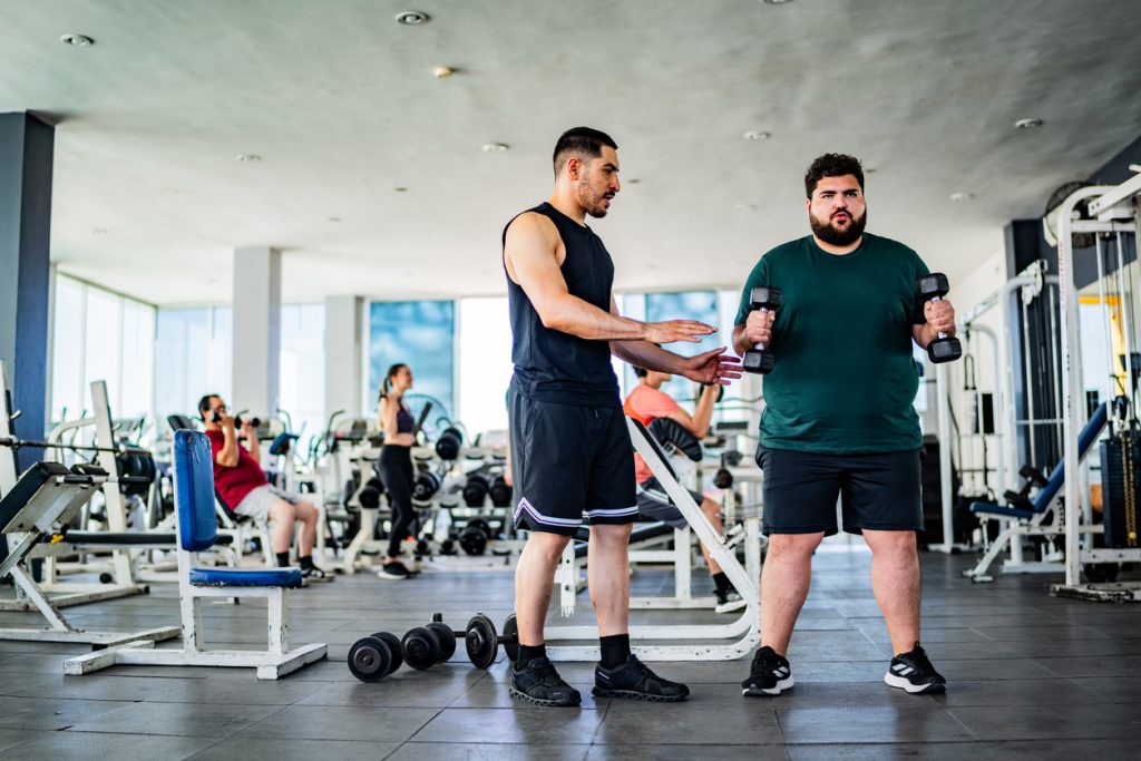 Homem levantando halteres em uma academia sob a supervisão de um instrutor físico, representando a mudança de hábitos e a prática de atividades necessárias para reverter o quadro de pré-diabetes.