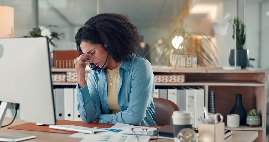 Mulher no escritório estressada com a mão no rosto, apoiada em sua mesa de trabalho e em frente ao computador.