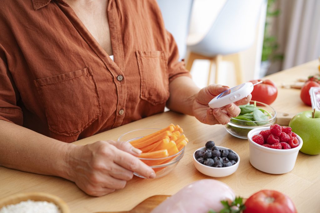 Mulher de blusa laranja sentada à mesa medindo o nível de açúcar no sangue com um glicosímetro. Ao redor dela há opções saudáveis que compõem uma dieta para diabetes, como potes com palitos de cenoura, mirtilos, framboesas, além de uma maçã verde e folhas de espinafre.