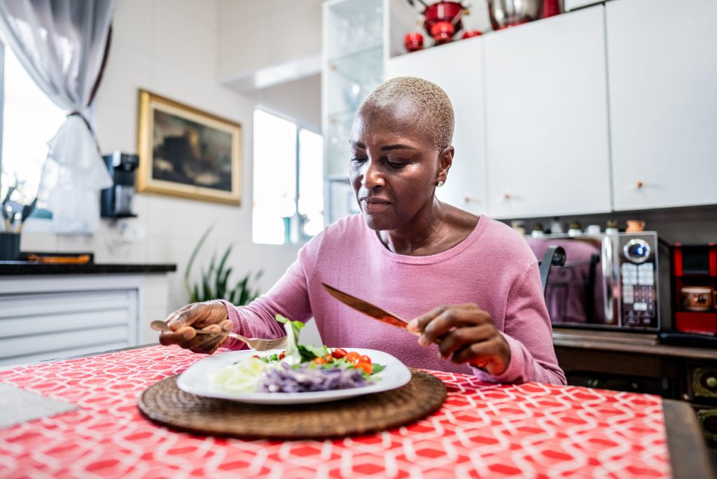 Mulher negra de cabelos curtos grisalhos sentada à mesa da sala de jantar, segurando garfo e faca enquanto se prepara para comer um prato de salada colorida com folhas verdes, tomate e repolho roxo. A imagem representa a adoção de estratégias alimentares leves para facilitar o trabalho do sistema digestório e aliviar a sensação de estômago cheio.