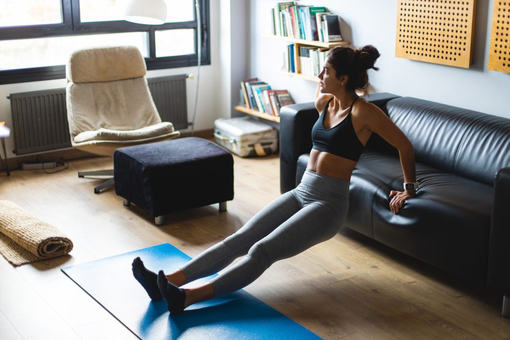 Mulher focada realizando exercício de tríceps apoiada em um sofá preto na sala de estar, comprovando que, com dedicação e adaptação do ambiente, a prática de exercícios para fazer em casa traz excelentes resultados para o corpo.