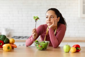 Mulher com expressão desanimada e uma fita métrica no pescoço, segurando um garfo com salada à frente de uma mesa com vegetais, ilustrando a falta de apetite comum em tratamentos de emagrecimento.