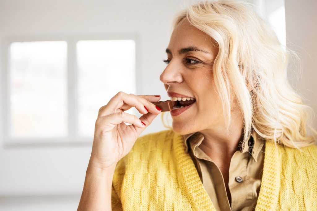 Mulher loira sorrindo e comendo um pedaço de chocolate, representando o apetite e a vontade de comer estimulados pela grelina.