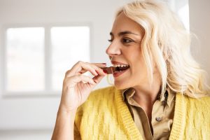 Mulher loira sorrindo e comendo um pedaço de chocolate, representando o apetite e a vontade de comer estimulados pela grelina.