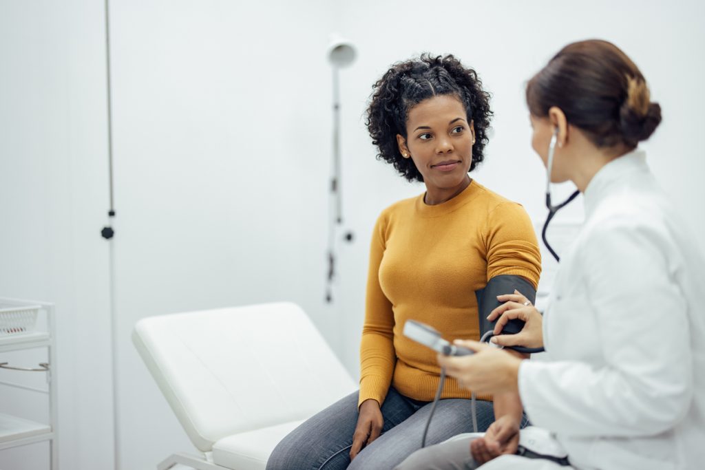 Uma médica de jaleco branco utiliza um esfigmomanômetro para medir a pressão arterial no braço de uma paciente mulher negra, que veste uma blusa amarela. Elas estão em um consultório médico claro e moderno. A imagem ilustra o diagnóstico e o monitoramento da pressão alta, um dos principais fatores de risco para doenças cardiovasculares.