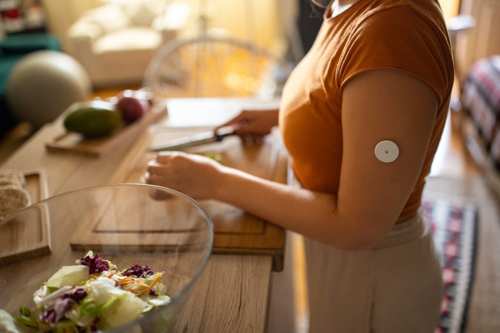 Mulher preparando alimentos em uma tábua de corte na cozinha. Ela usa um sensor de monitoramento contínuo de glicose no braço esquerdo. Em primeiro plano, há uma tigela de vidro com uma salada de folhas variadas, destacando os melhores alimentos para incluir na dieta para diabetes.