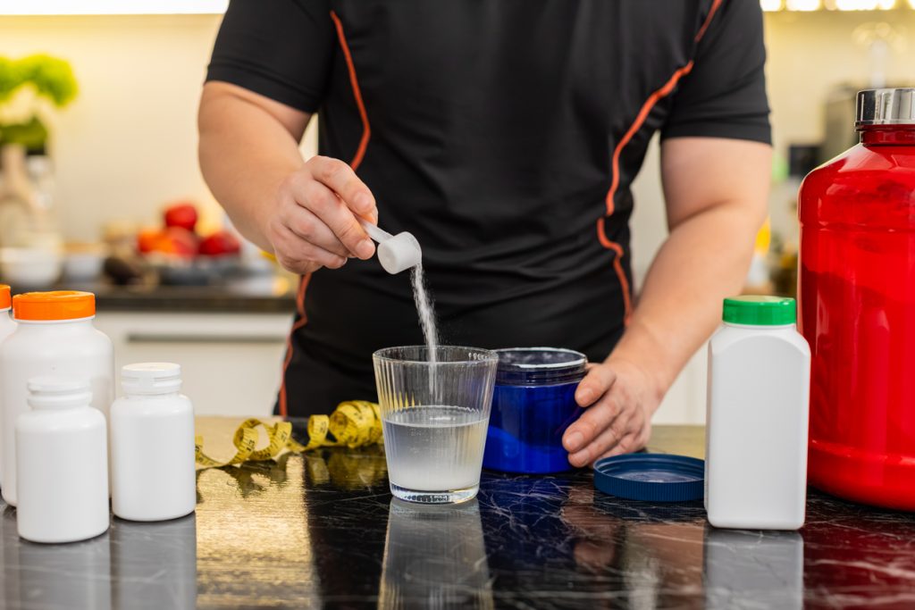 Homem preparando sua dose de creatina em uma bancada com outros suplementos em volta. Ele usa roupas leves para exercícios físicos.