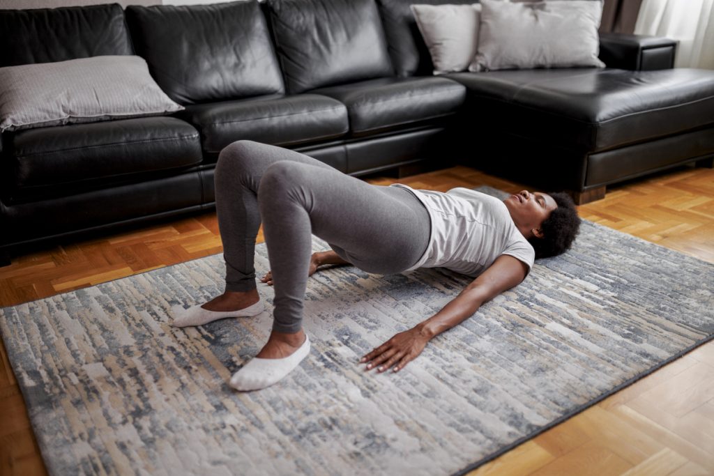 Mulher negra deitada de barriga para cima em um tapete na sala, elevando o quadril na posição de ponte de glúteos. Uma ótima opção para incluir na sua rotina de exercícios para fazer em casa.
