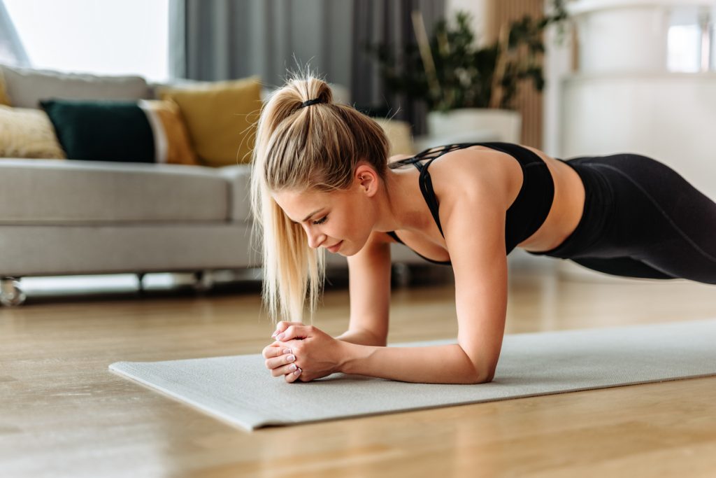 Mulher loira com roupa de ginástica preta sustentando o peso do corpo na posição de prancha abdominal com os antebraços apoiados em um tapete de yoga, ideal para fortalecer o core com exercícios para fazer em casa.