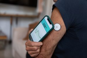 Close-up de uma pessoa utilizando um sensor de monitoramento contínuo de glicose no braço e verificando os níveis de açúcar no sangue através de um smartphone.