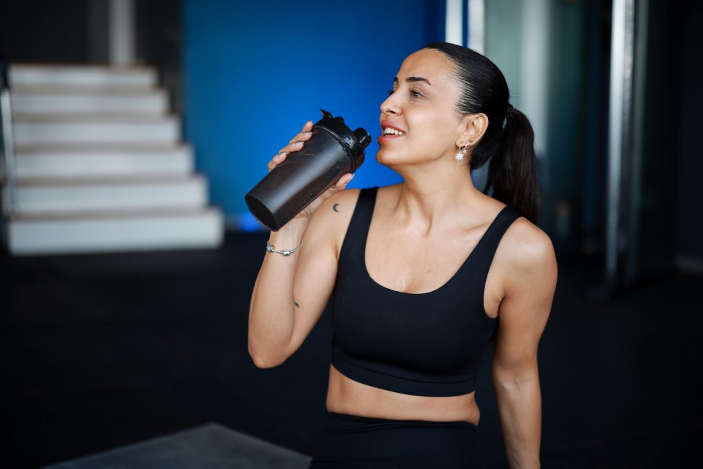 Mulher sorridente com roupas de ginástica pretas fazendo uma pausa na academia e bebendo o suplemento em uma coqueteleira preta. A foto ilustra a seção sobre os diferentes perfis de pessoas que podem usar e para que serve whey protein.