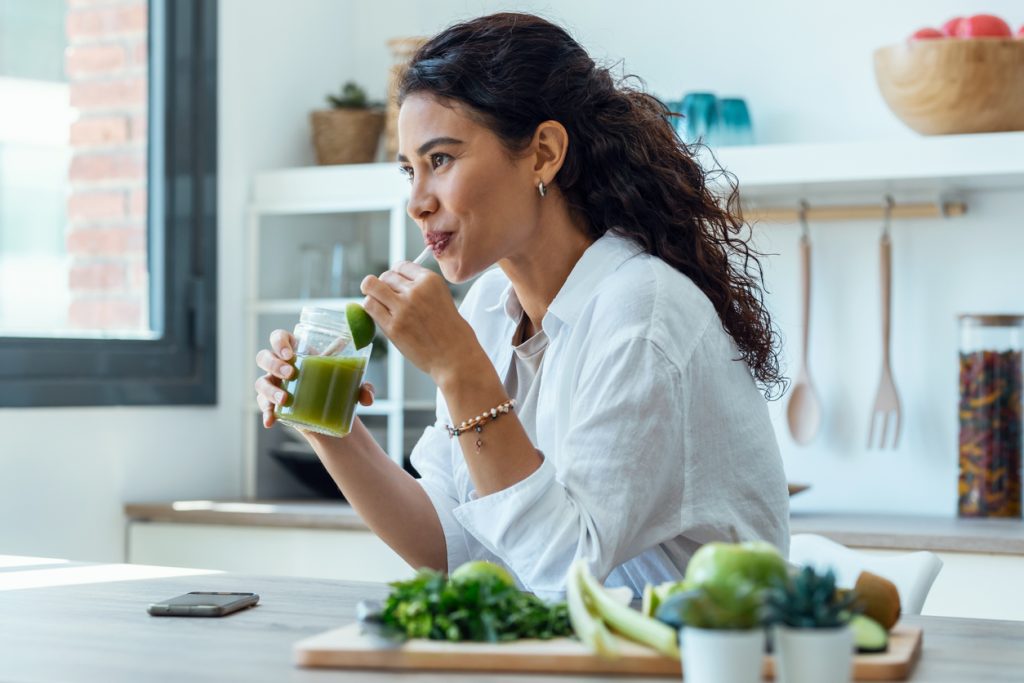 Mulher sorridente tomando um suco verde com canudo em uma cozinha iluminada, com vegetais frescos sobre a mesa. A imagem ilustra o estilo de vida saudável associado ao equilíbrio da leptina, o hormônio da saciedade.