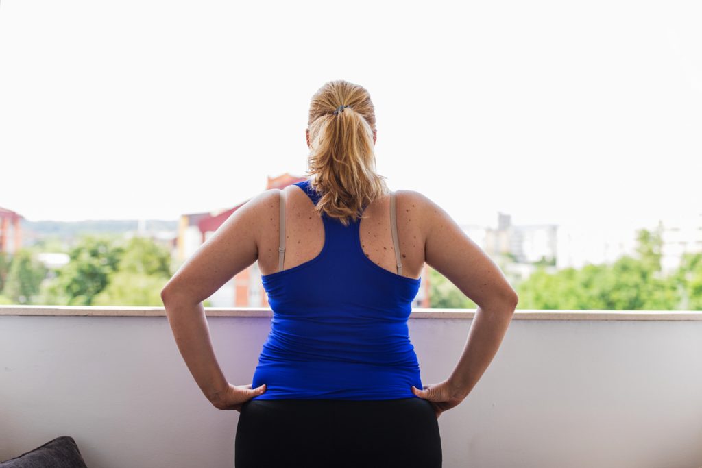 Mulher vista de costas usando uma blusa azul de ginástica e com as mãos na cintura. Níveis altos de grelina aumentam o apetite e podem contribuir para o ganho de peso.