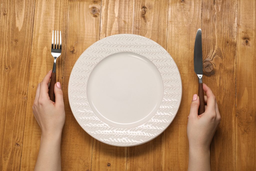 Visão superior de mãos femininas segurando um garfo e uma faca ao lado de um prato branco vazio sobre uma mesa de madeira, simbolizando a redução da fome e o controle da grelina.