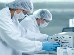 Dois profissionais paramentados com jaleco branco, touca, máscara e luvas azuis operam um equipamento industrial durante a produção de SEMAVY, caneta de semaglutida sintética aprovada pela Anvisa.