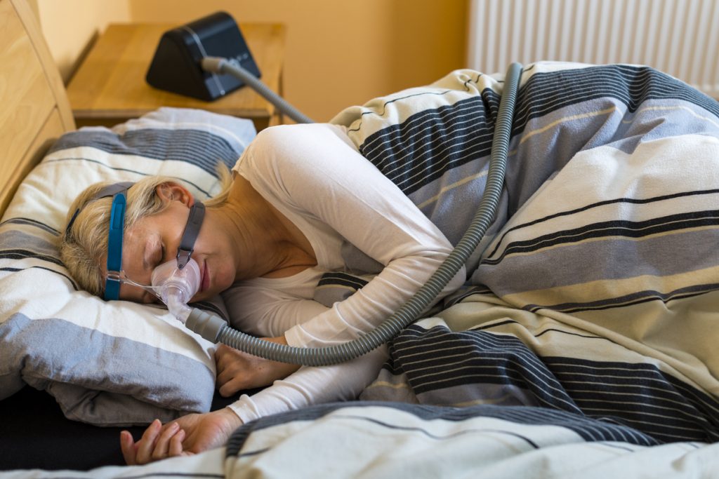 Mulher loira dormindo de lado na cama coberta por um edredom, utilizando uma máscara facial de CPAP conectada por um tubo ao aparelho na mesa de cabeceira, que é o tratamento padrão para apneia do sono.