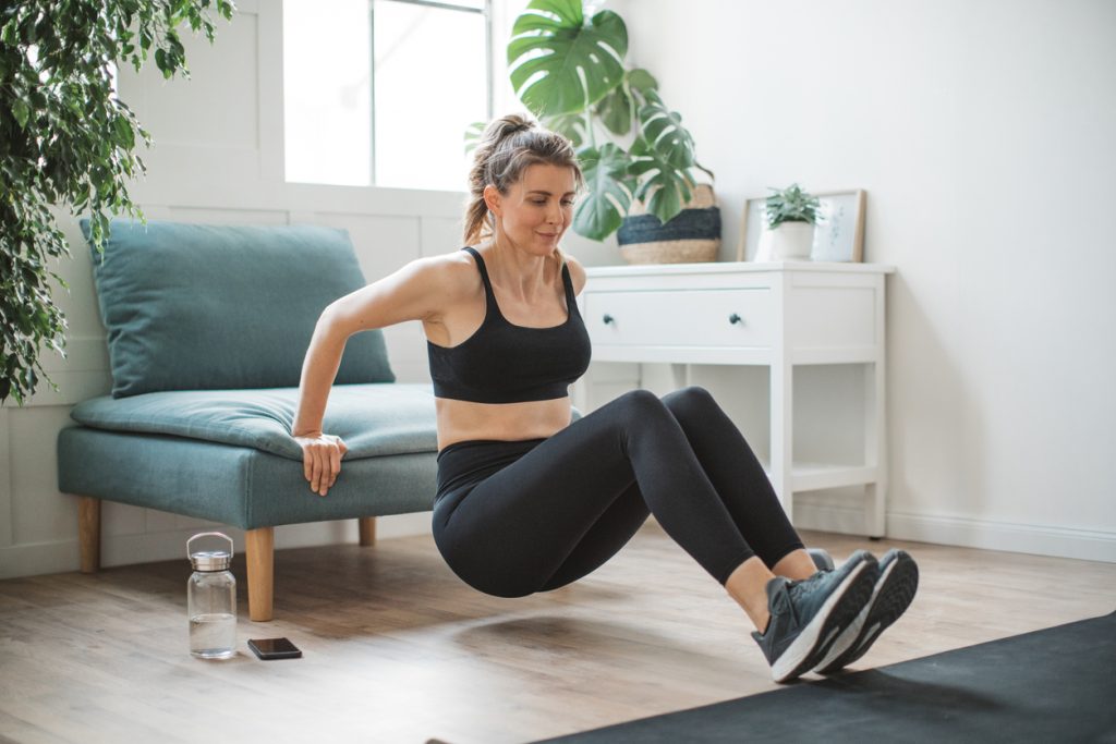 Mulher loira com roupas de ginástica pretas fazendo o exercício de tríceps apoiando as mãos na beirada de um pufe/sofá verde na sala. Uma forma prática de adaptar exercícios para fazer em casa utilizando os móveis.