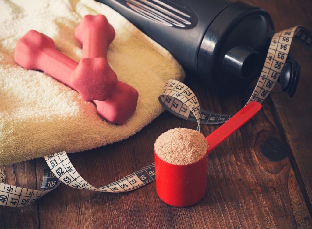 Scoop de whey protein ao lado de fita métrica, copo e halteres de academia.