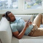 Fotografia de um homem negro deitado confortavelmente em um sofá claro próximo a uma grande janela. Ele veste uma camiseta azul claro, está usando fones de ouvido e focado na tela do seu smartphone. A imagem ilustra o comportamento inativo associado ao sedentarismo, tema central do artigo sobre emagrecimento.
