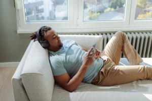 Fotografia de um homem negro deitado confortavelmente em um sofá claro próximo a uma grande janela. Ele veste uma camiseta azul claro, está usando fones de ouvido e focado na tela do seu smartphone. A imagem ilustra o comportamento inativo associado ao sedentarismo, tema central do artigo sobre emagrecimento.