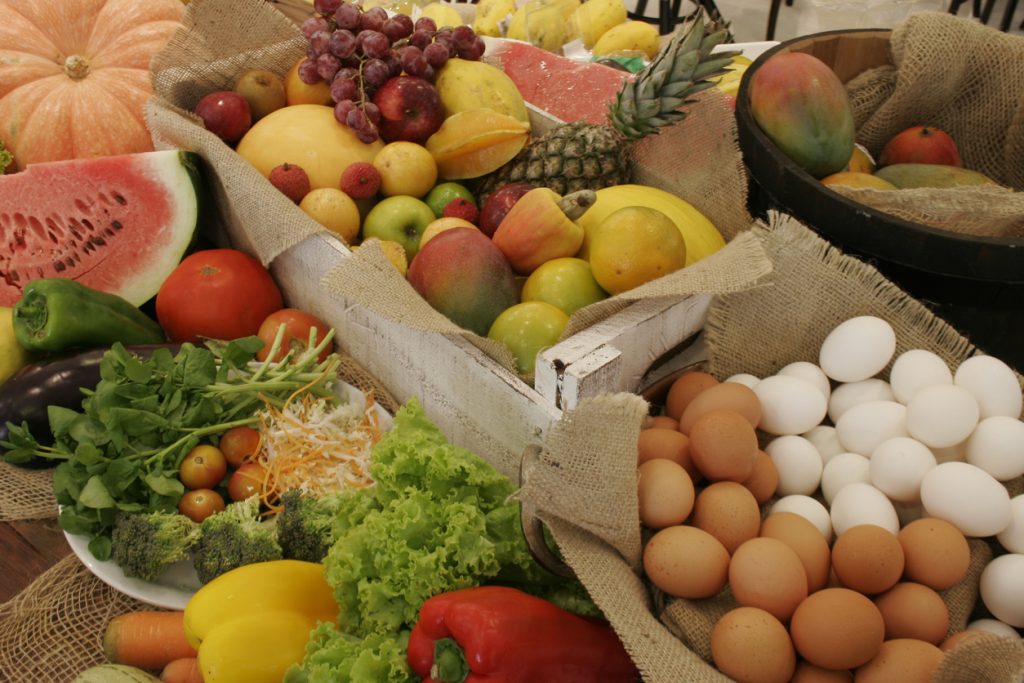 Exibição rústica de uma abundância de alimentos naturais inteiros, incluindo caixotes de frutas tropicais misturadas (manga, uva, abacaxi), vegetais frescos e ovos de feira, representando o alicerce nutricional contra alimentos ultraprocessados.