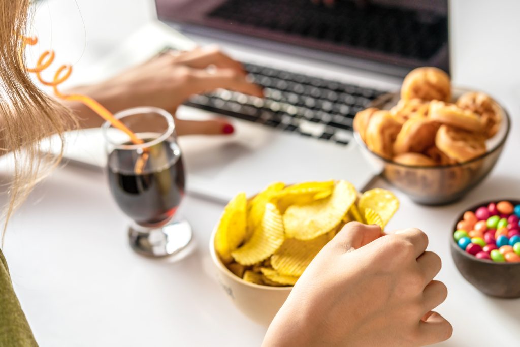 Visão aproximada de uma pessoa utilizando um notebook enquanto consome diversos snacks ultraprocessados, como salgadinhos, doces e refrigerante, representando a alimentação emocional causada pela ansiedade.