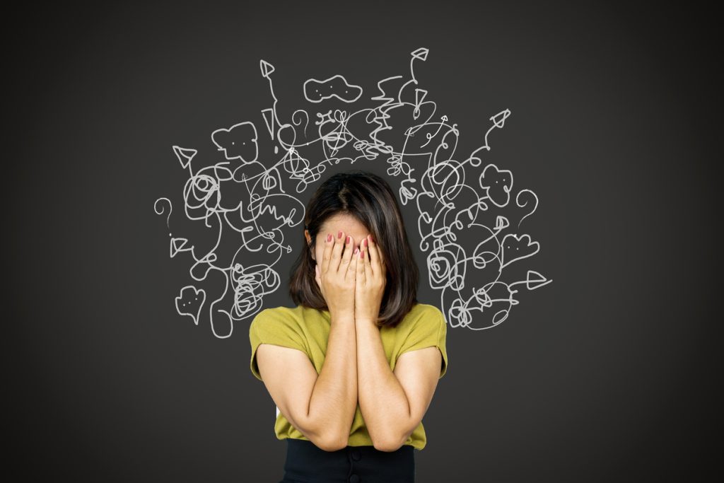 Uma mulher com as mãos cobrindo o rosto em sinal de frustração, com ilustrações de rabiscos e setas confusas ao redor de sua cabeça, simbolizando a sobrecarga mental e a ansiedade.