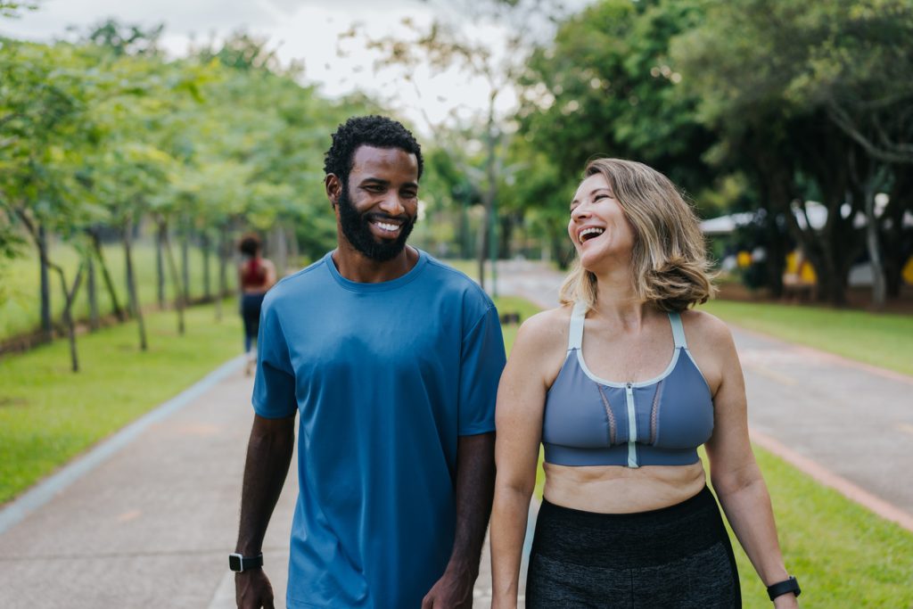 Homem e mulher vestindo roupas esportivas caminhando lado a lado e sorrindo em uma pista de parque arborizado, representando a adoção de hábitos saudáveis e exercícios físicos para prevenir os sintomas de AVC no dia a dia.
