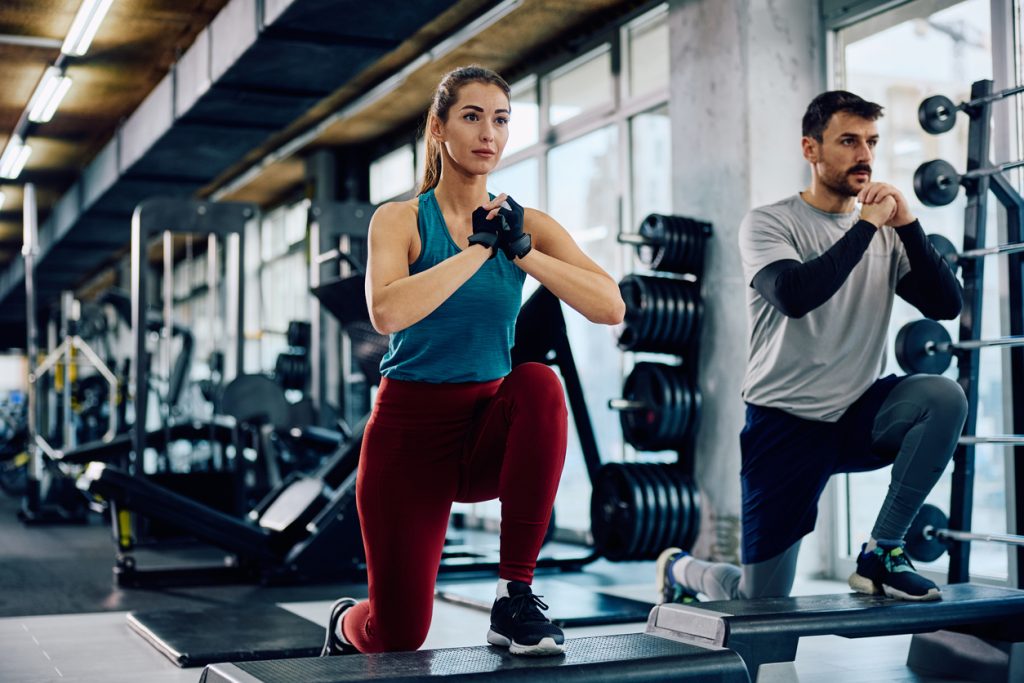 Uma mulher e um homem realizando exercício de afundo com uma perna apoiada no step em uma academia. Ter consistência nos treinos e exercícios práticos é a melhor forma de garantir que a musculação emagrece no dia a dia.