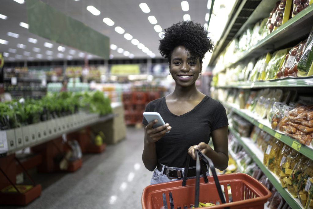 Jovem mulher sorridente em um supermercado, segurando uma cesta de compras e seu smartphone, focada na seção de hortifrúti com vegetais frescos, buscando dicas para manter uma alimentação balanceada livre de alimentos ultraprocessados.