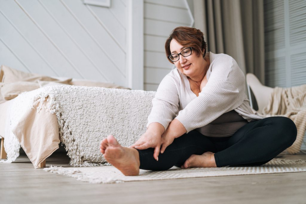 Mulher com sobrepeso usando óculos, sentada no chão de um quarto aconchegante, sorrindo e com expressão serena enquanto se inclina para a frente fazendo exercícios de alongamento na perna direita, representando a recuperação da mobilidade e o tratamento ativo para a osteoartrite.