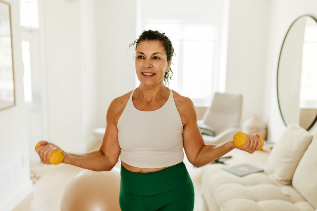 Uma mulher de meia-idade sorrindo enquanto faz exercícios de bíceps com pequenos halteres amarelos em sua sala de estar. Uma imagem focada na estratégia de atividade física para gerenciar o peso e a massa muscular na menopausa.