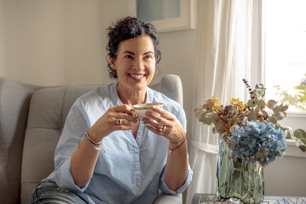 Uma mulher madura com cabelos cacheados e expressão serena, segurando uma xícara de chá perto de um buquê de flores secas e azuis. Representa uma estratégia de autocuidado e bem-estar para lidar com os sintomas da menopausa.