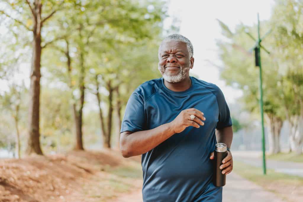 Homem mais velho, com barba grisalha e roupas esportivas, correndo sorridente em um parque arborizado e segurando uma garrafa térmica. Imagem de capa para o artigo sobre como a andropausa pode impactar no peso do homem e na vida do casal.
